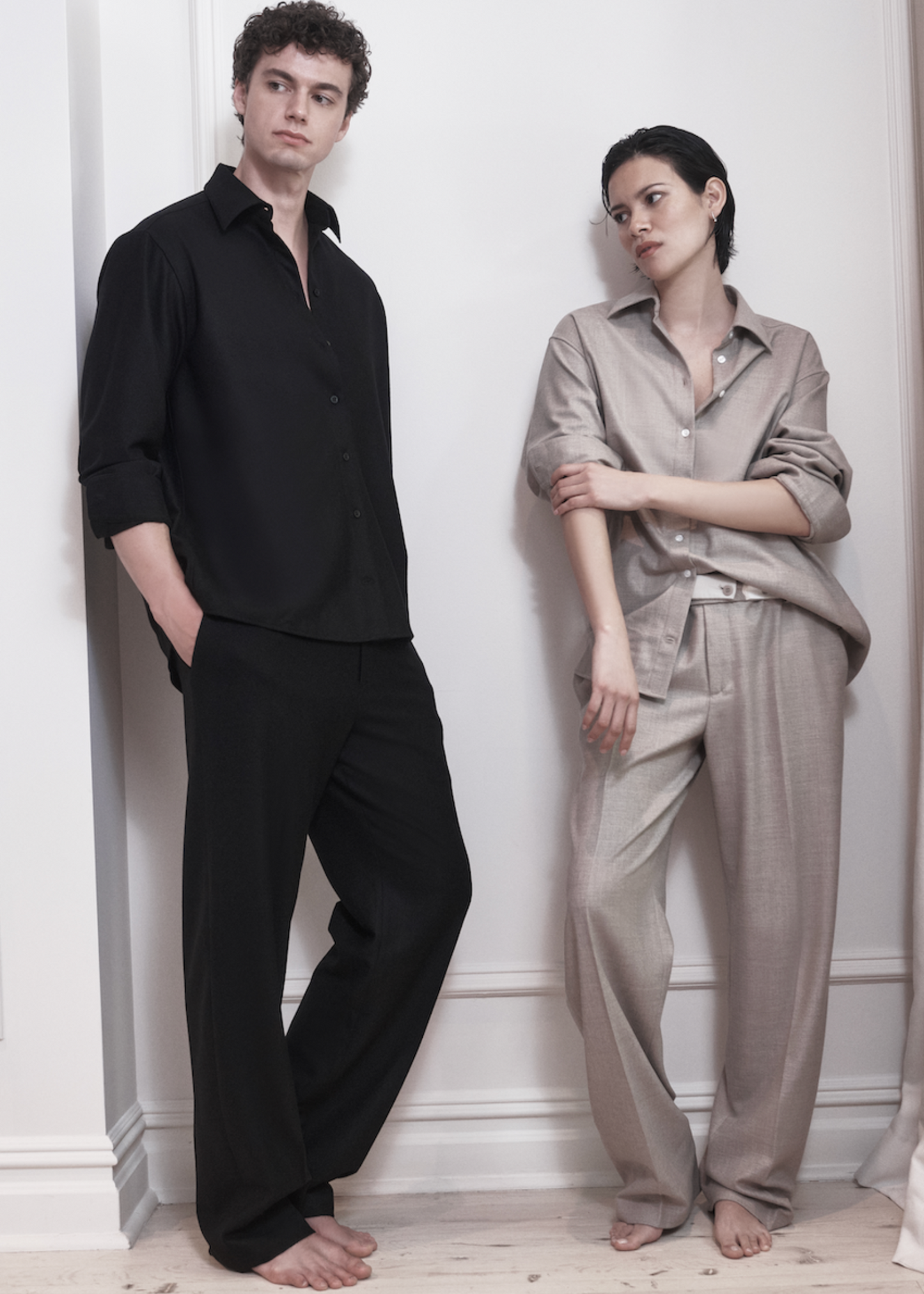 Two people wearing black and beige sets standing against a white wall.